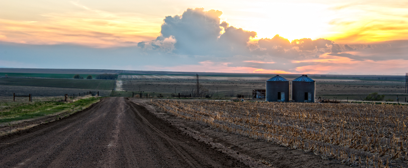 Rural landscape with dirt road, cornfield, silos, and dramatic sunset sky.