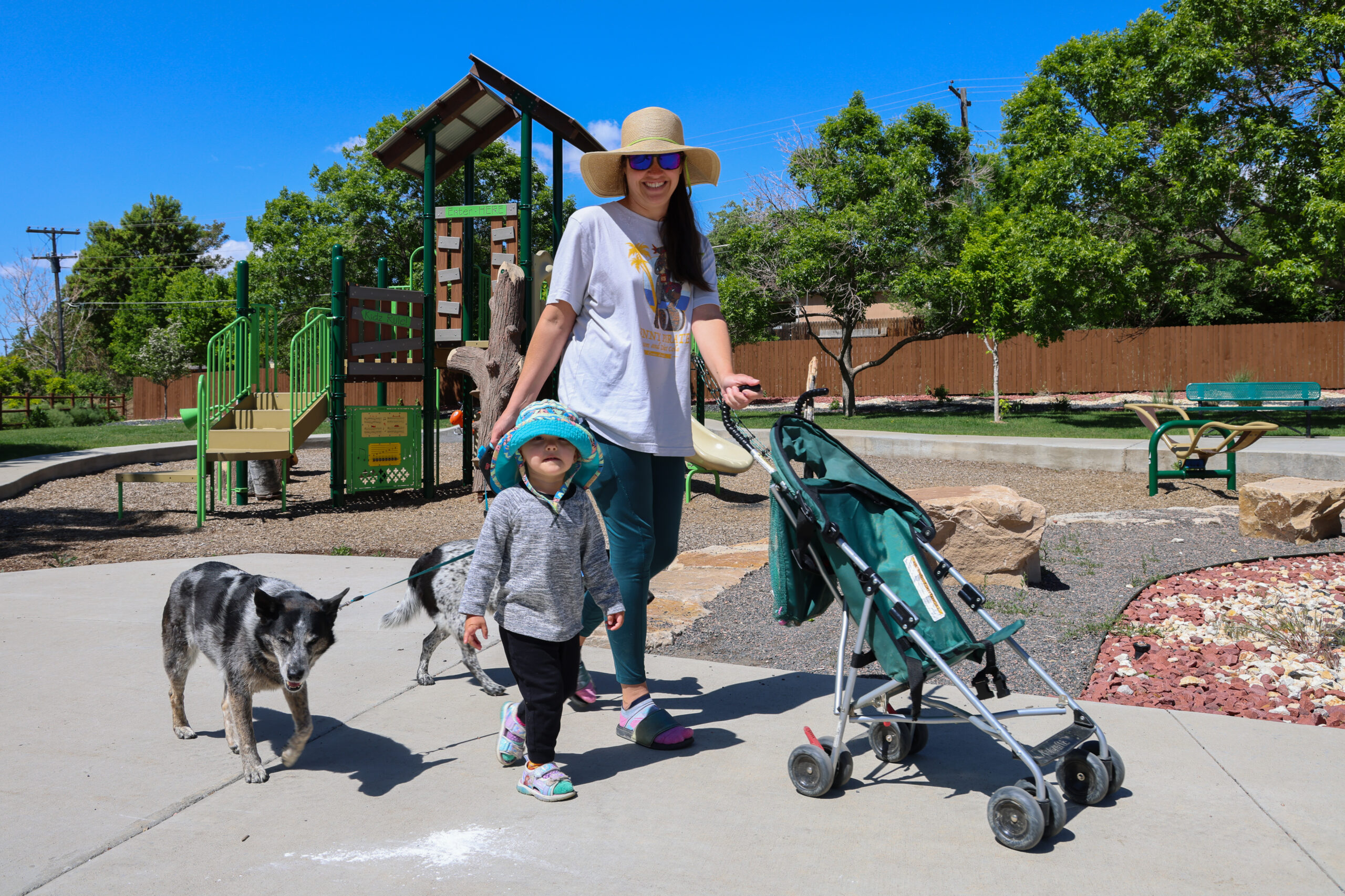 A woman in a sunhat smiles next to a child and stroller at a park, with a dog walking beside them.