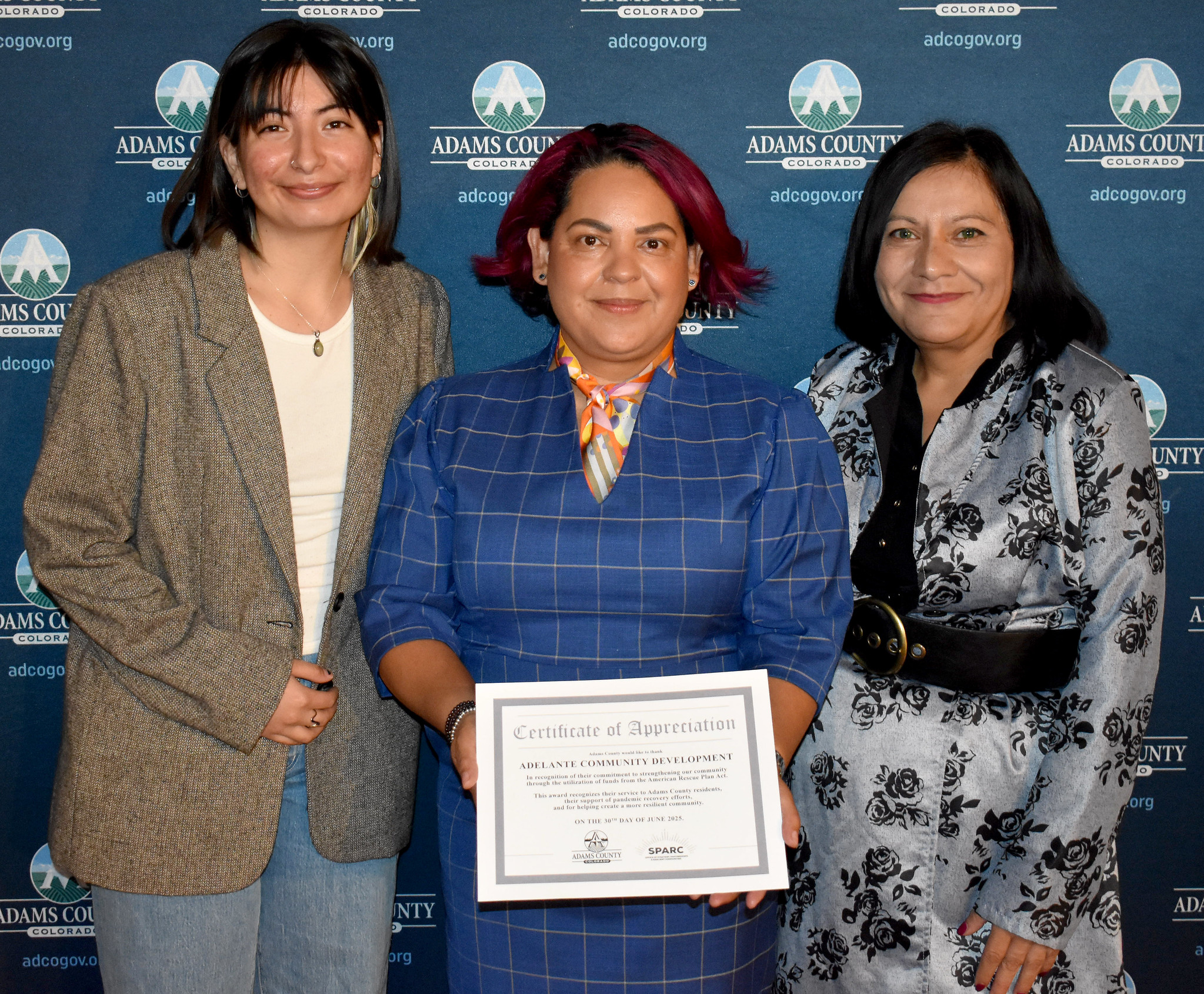 Three individuals smiling, one holding a "Certificate of Appreciation" for Adelante Community Development, with an Adams County backdrop.