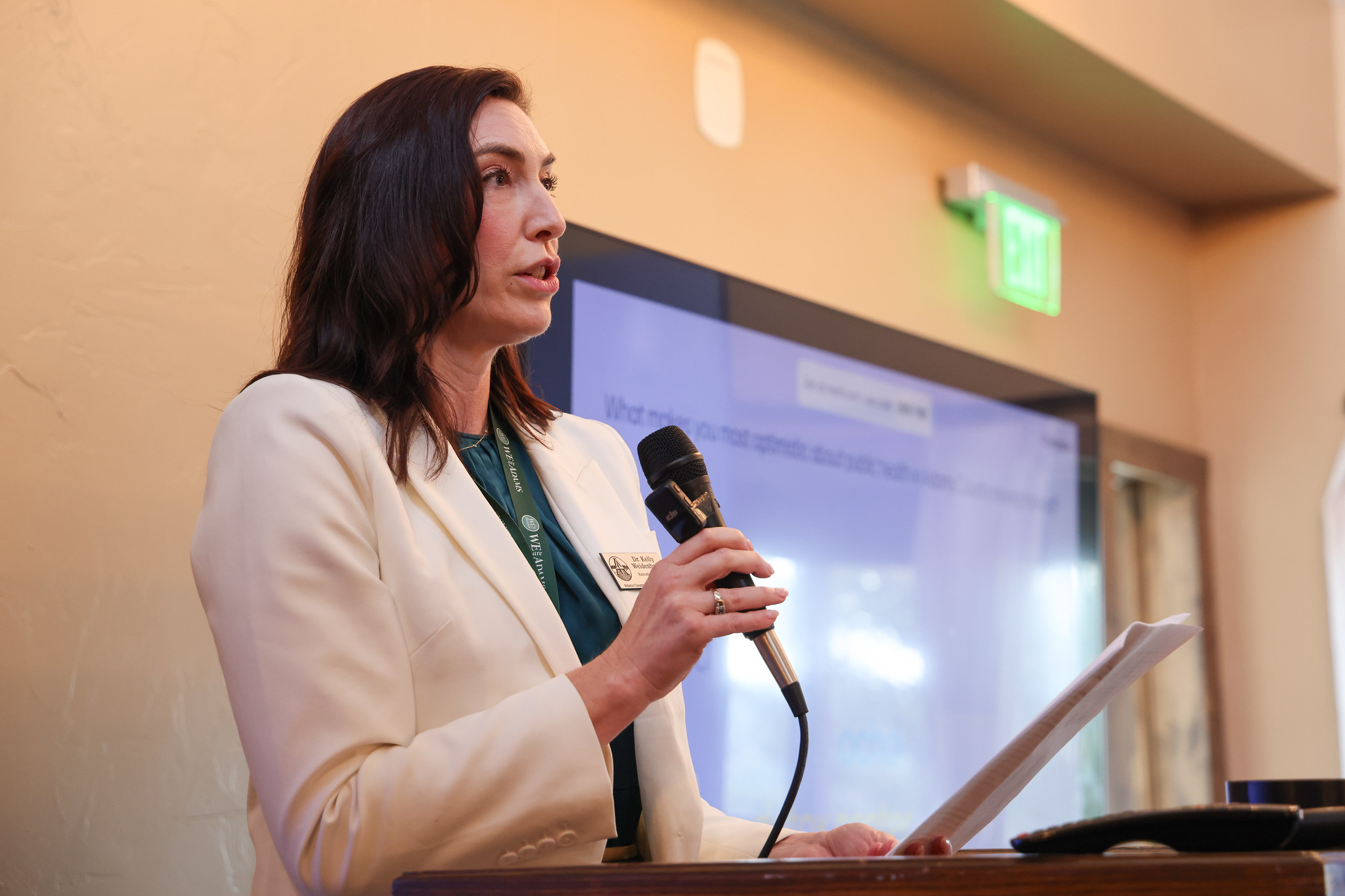 Dr. Kelley speaking at podium with microphone, wearing a nametag and white blazer.
