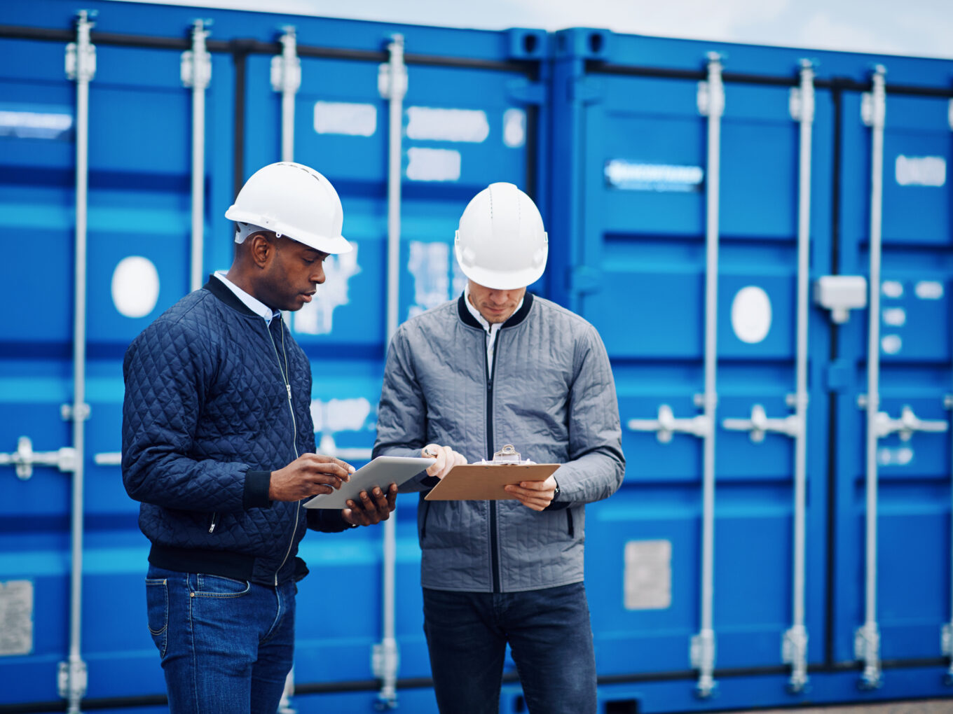 Two workers in hard hats discuss logistics near blue shipping containers.