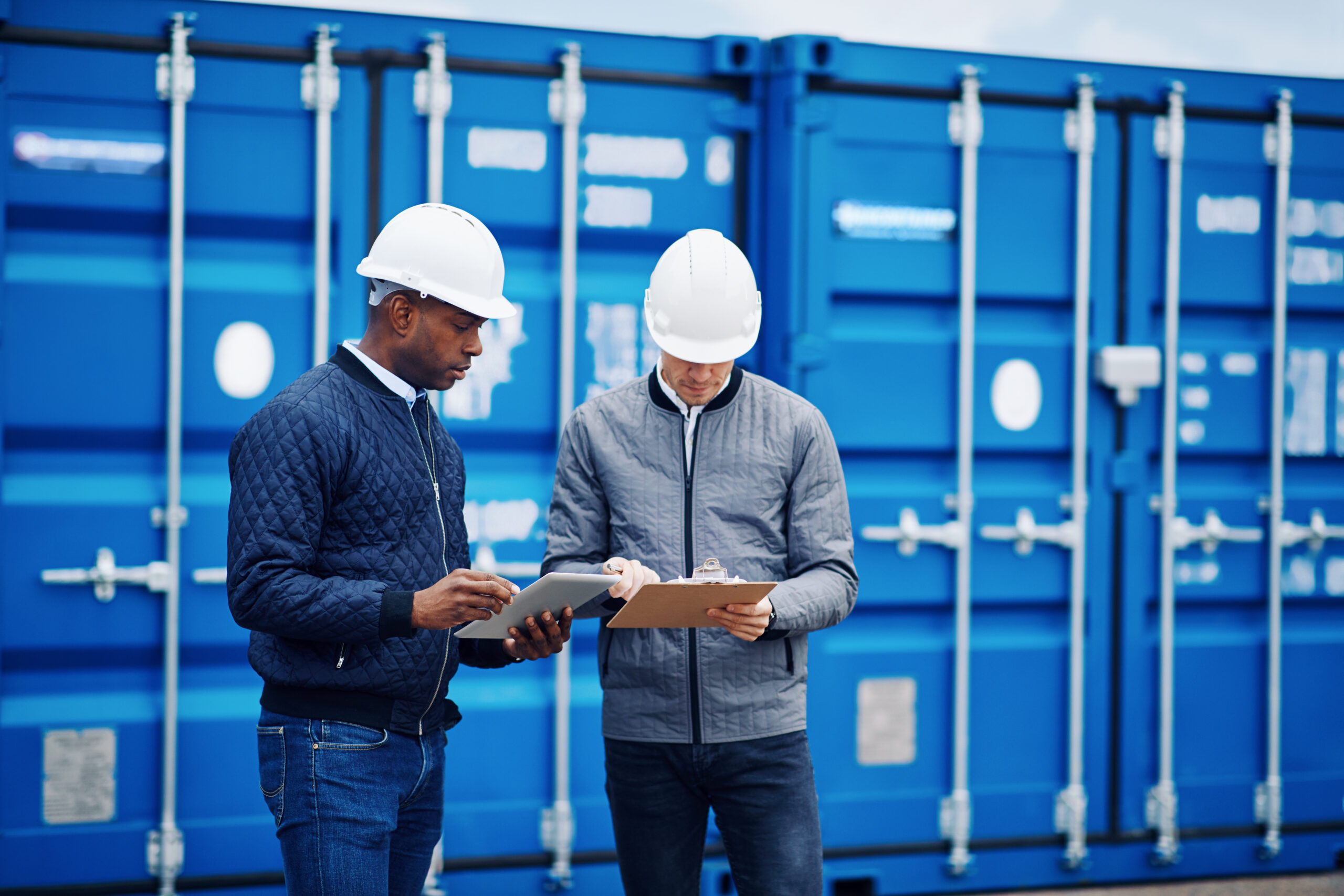 Two workers in hard hats discuss logistics near blue shipping containers.