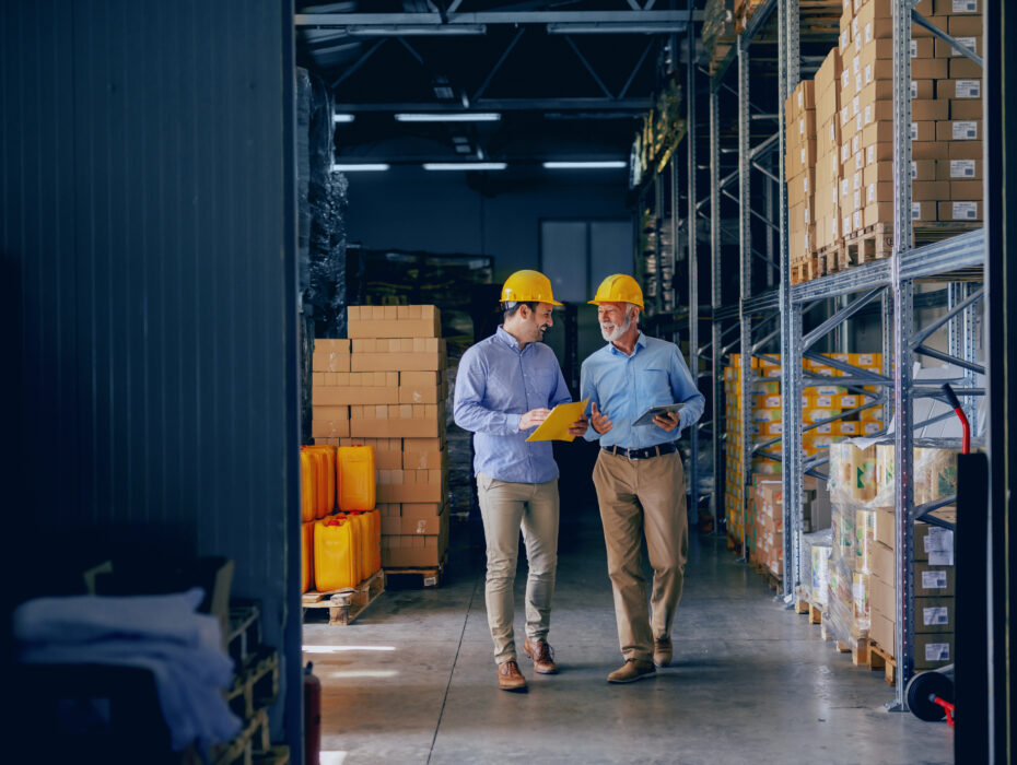 Two men in a warehouse discussing, both wearing safety helmets.