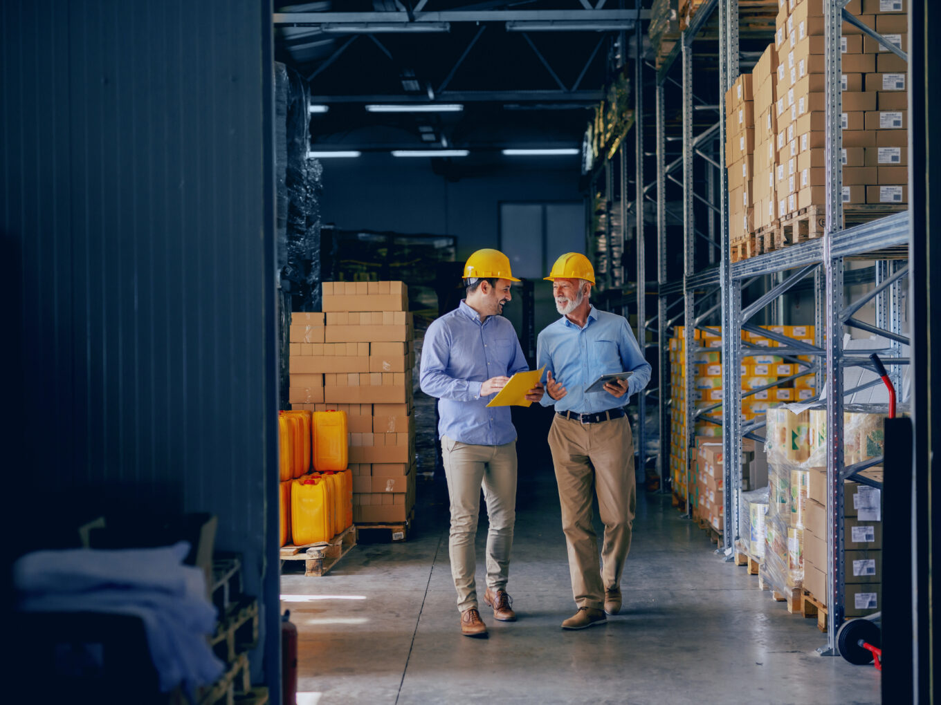 Two men in a warehouse discussing, both wearing safety helmets.
