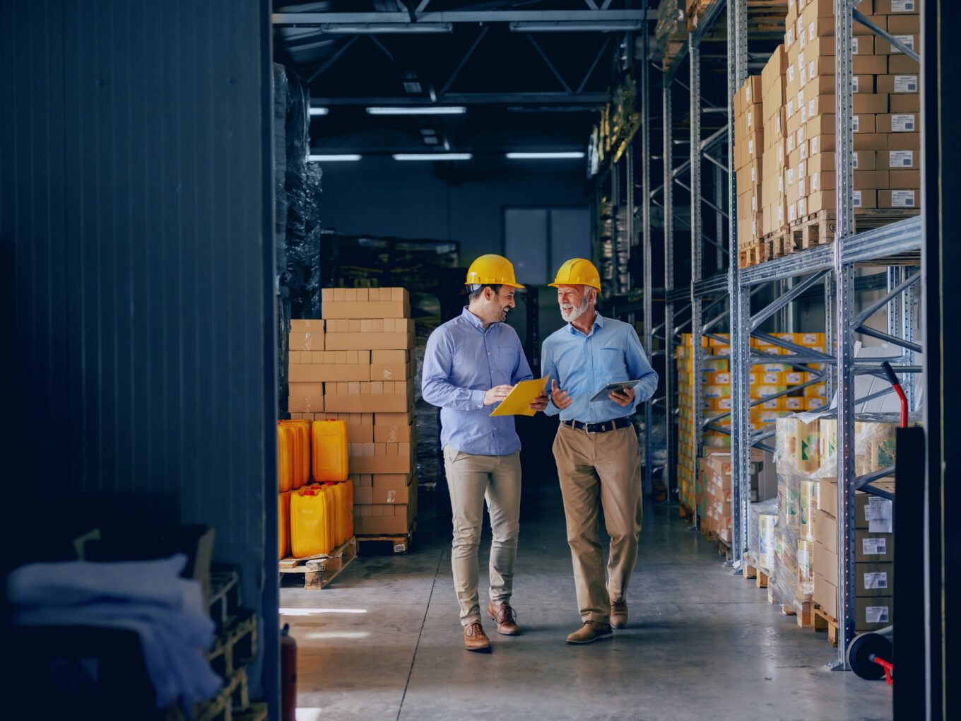 Two men in a warehouse discussing, both wearing safety helmets.