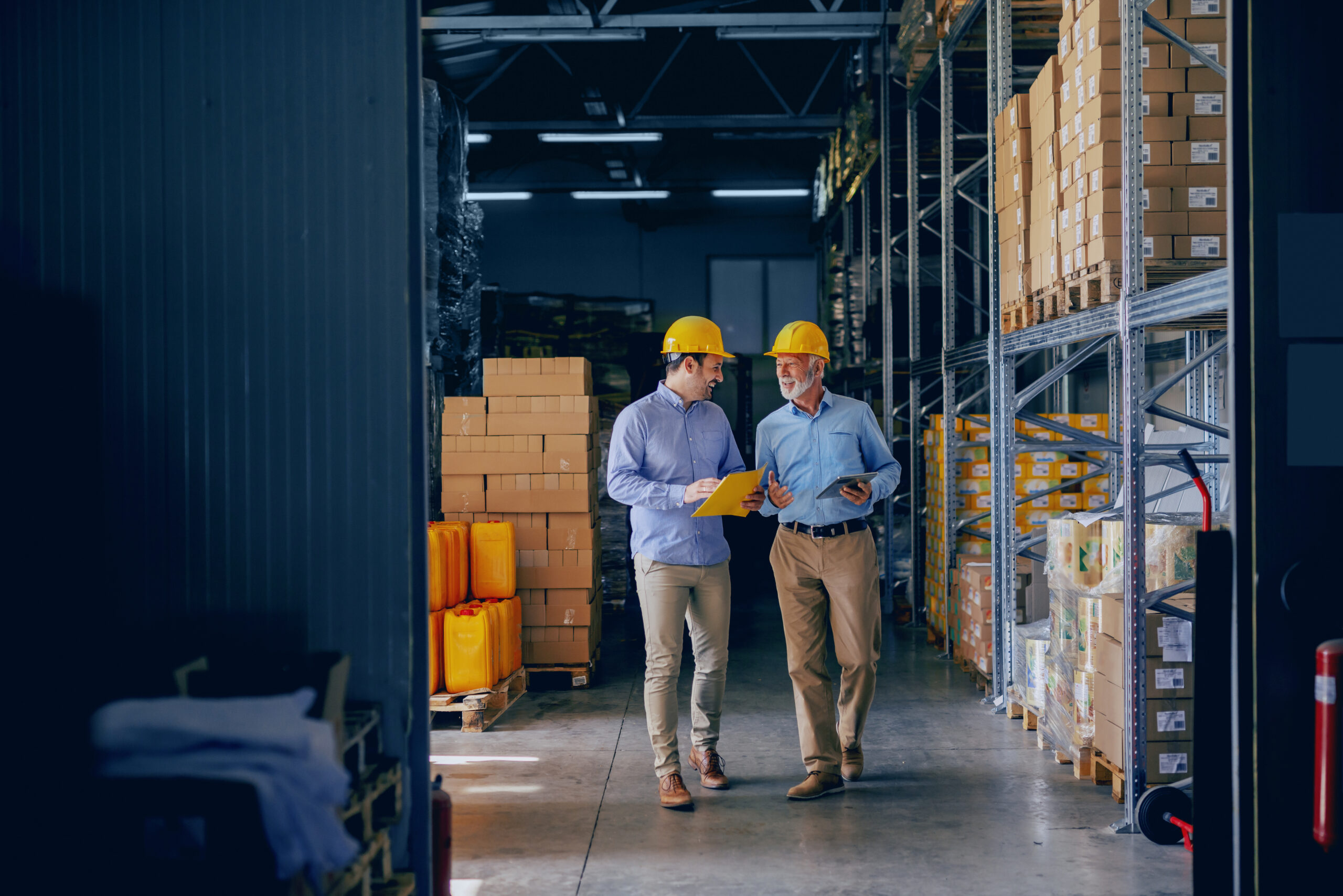 Two men in a warehouse discussing, both wearing safety helmets.