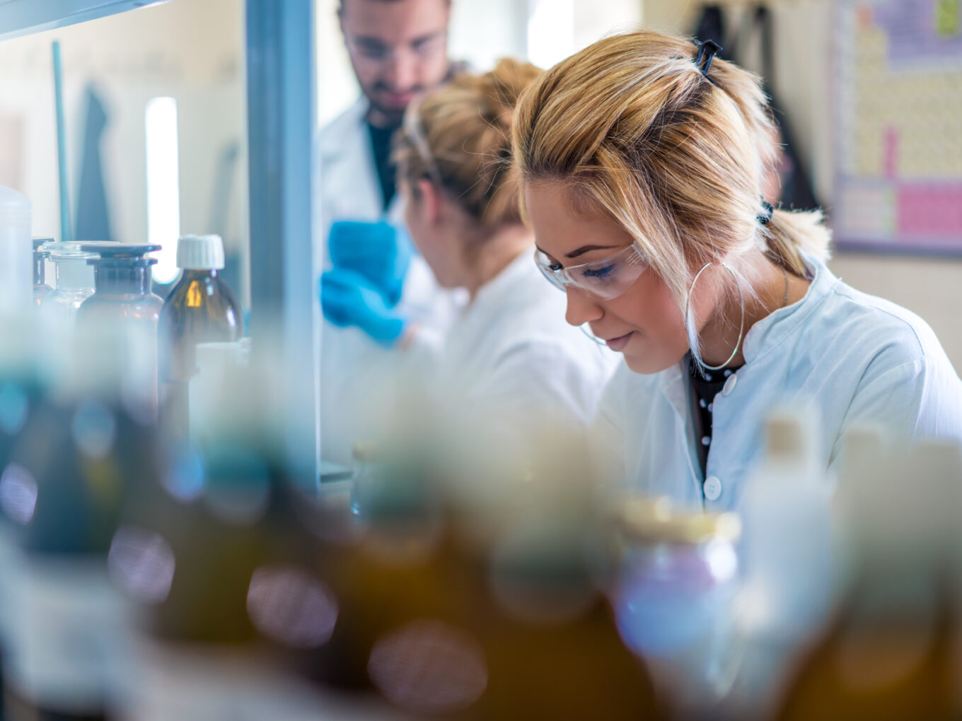 Woman in lab coat and goggles focuses intently, surrounded by bottles.