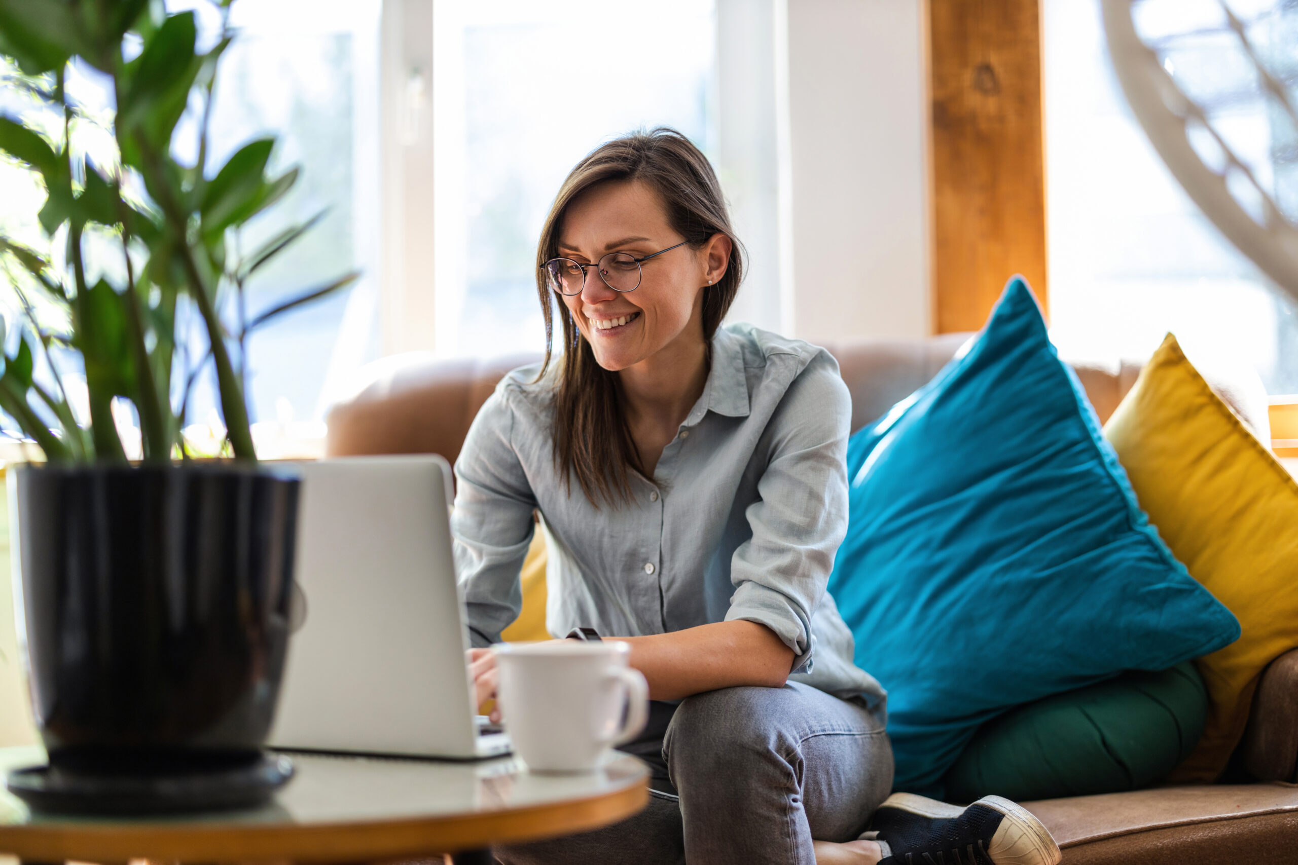 A person smiling while using a laptop on a couch, with colorful pillows and a plant nearby.