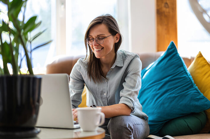 Person smiling at laptop in cozy, sunlit living room.