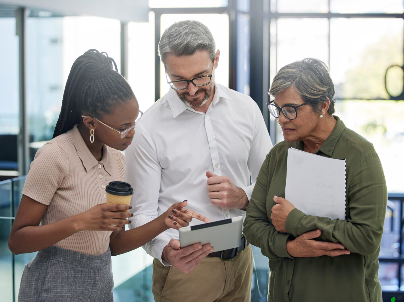 Three colleagues discuss a document on a tablet, one holding a coffee, another papers.