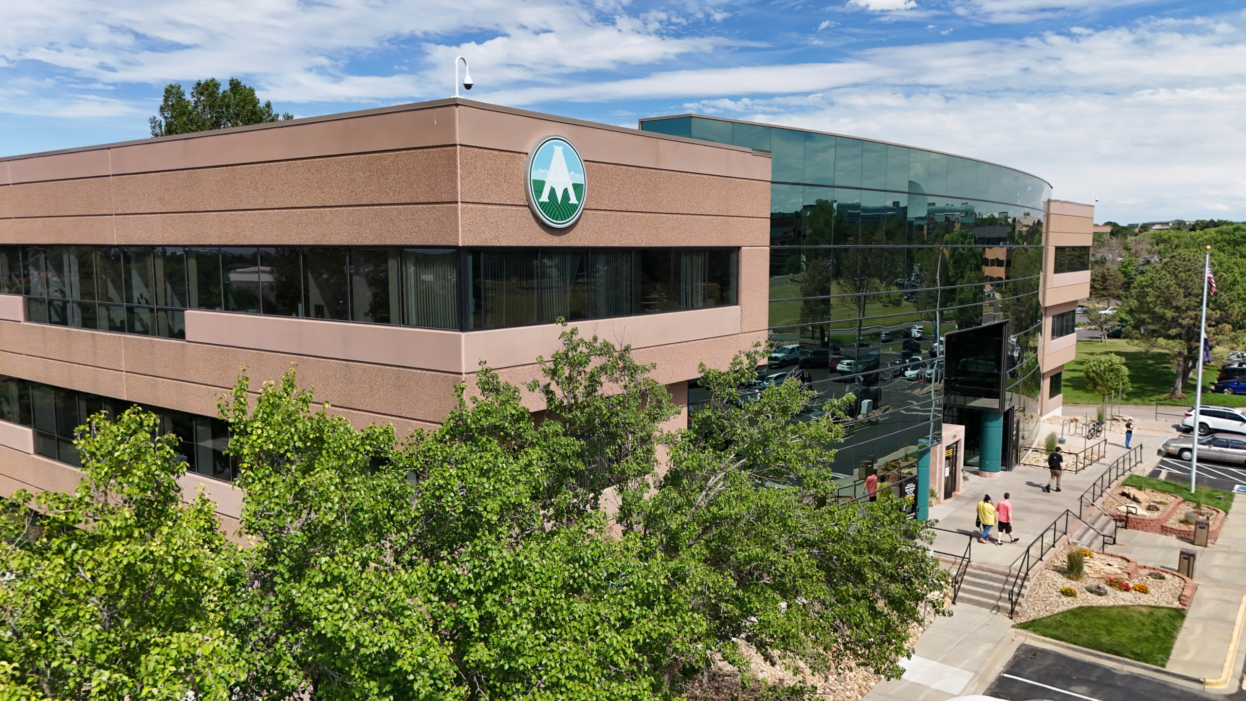 Modern office building with reflective glass facade and mountain logo, surrounded by trees and people walking nearby