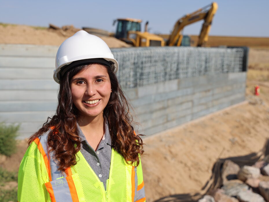 3V0A8027.jpg: Smiling construction worker in hard hat and safety vest at site with heavy machinery.