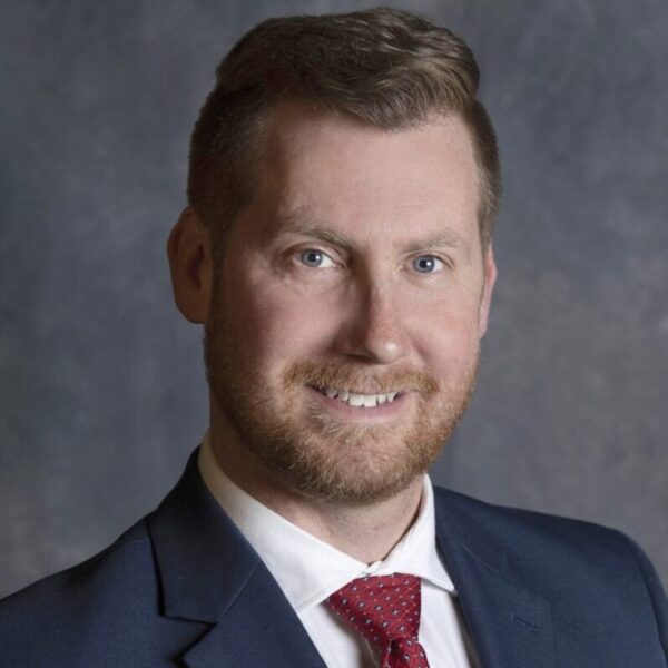 Aiken Bill smiling in formal attire with a blue suit and red tie