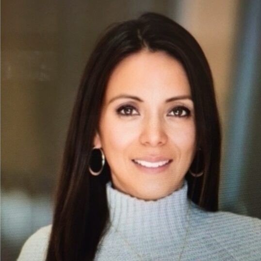 Delia Sanchez smiling, wearing a light blue turtleneck sweater and hoop earrings