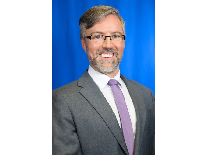 Ethan Rouse smiling man in glasses and suit with purple tie against blue background