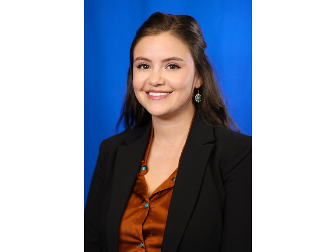 Marki Frazier smiling against a blue background, wearing a black blazer and brown shirt.