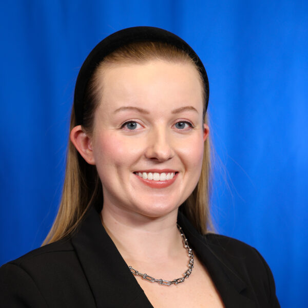 Victoria smiling in a black blazer against a blue background.